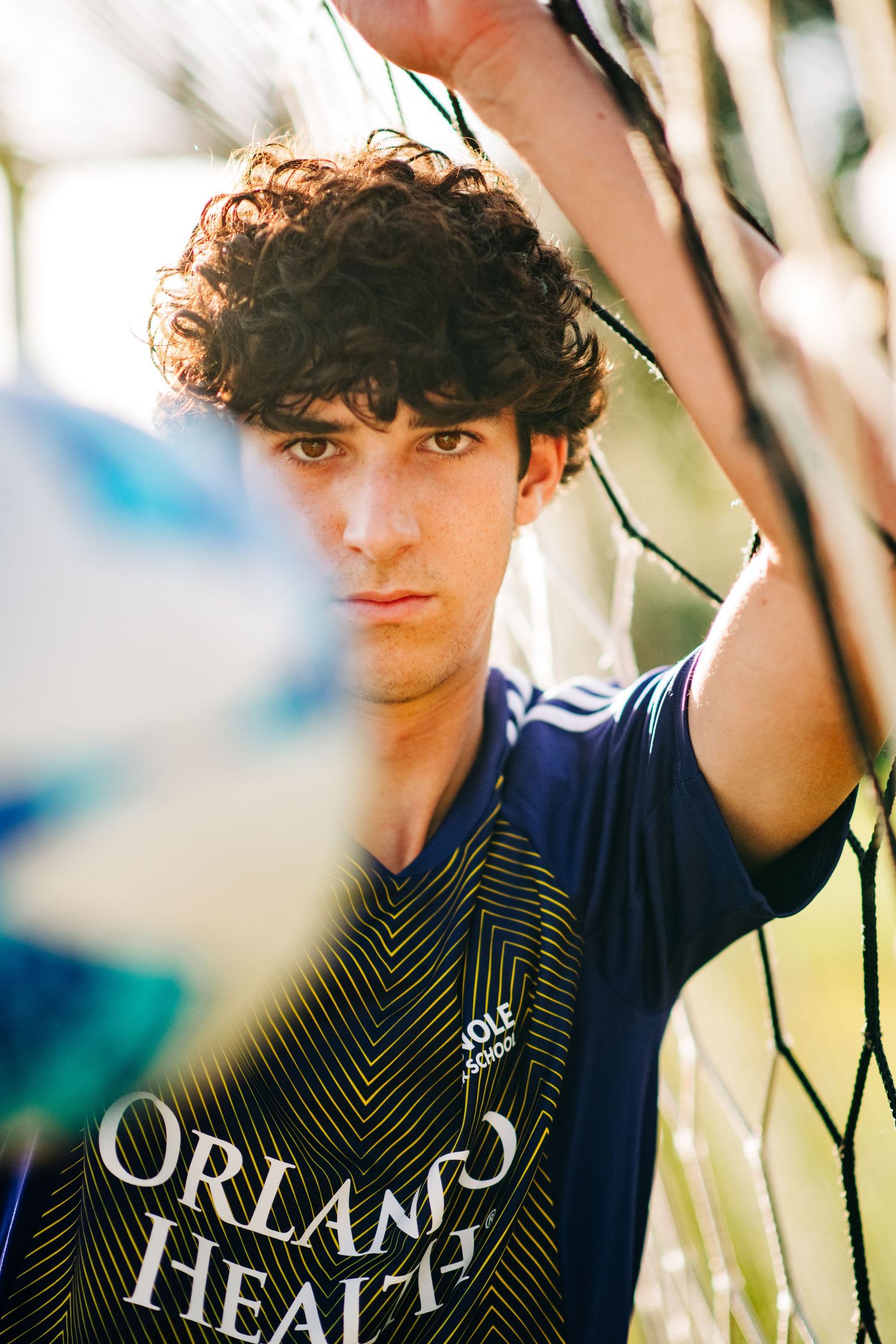 Young male soccer player in Orlando Health jersey leaning against goal net, holding netting overhead.