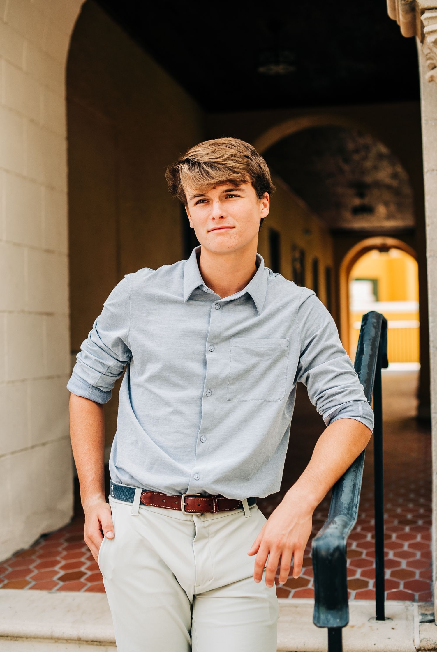 Young man in light blue shirt and khaki pants posing in an arched corridor for senior portraits.