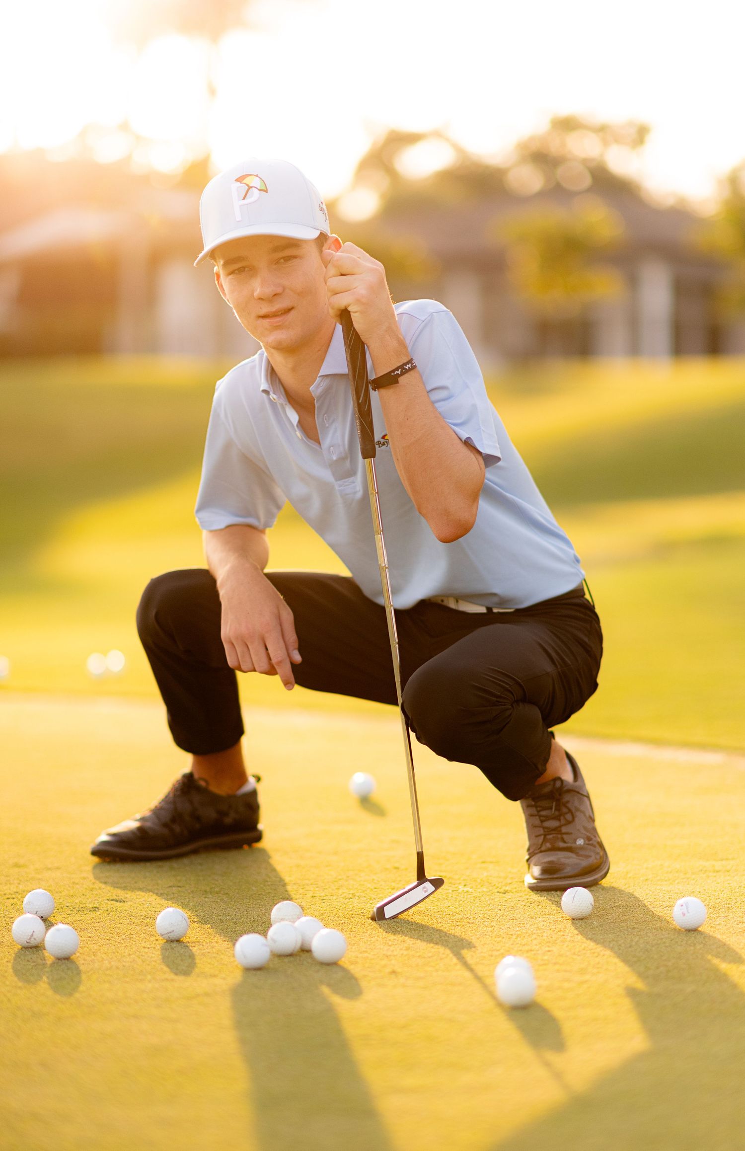 Teen golfer crouching on green with putter surrounded by golf balls at sunset.