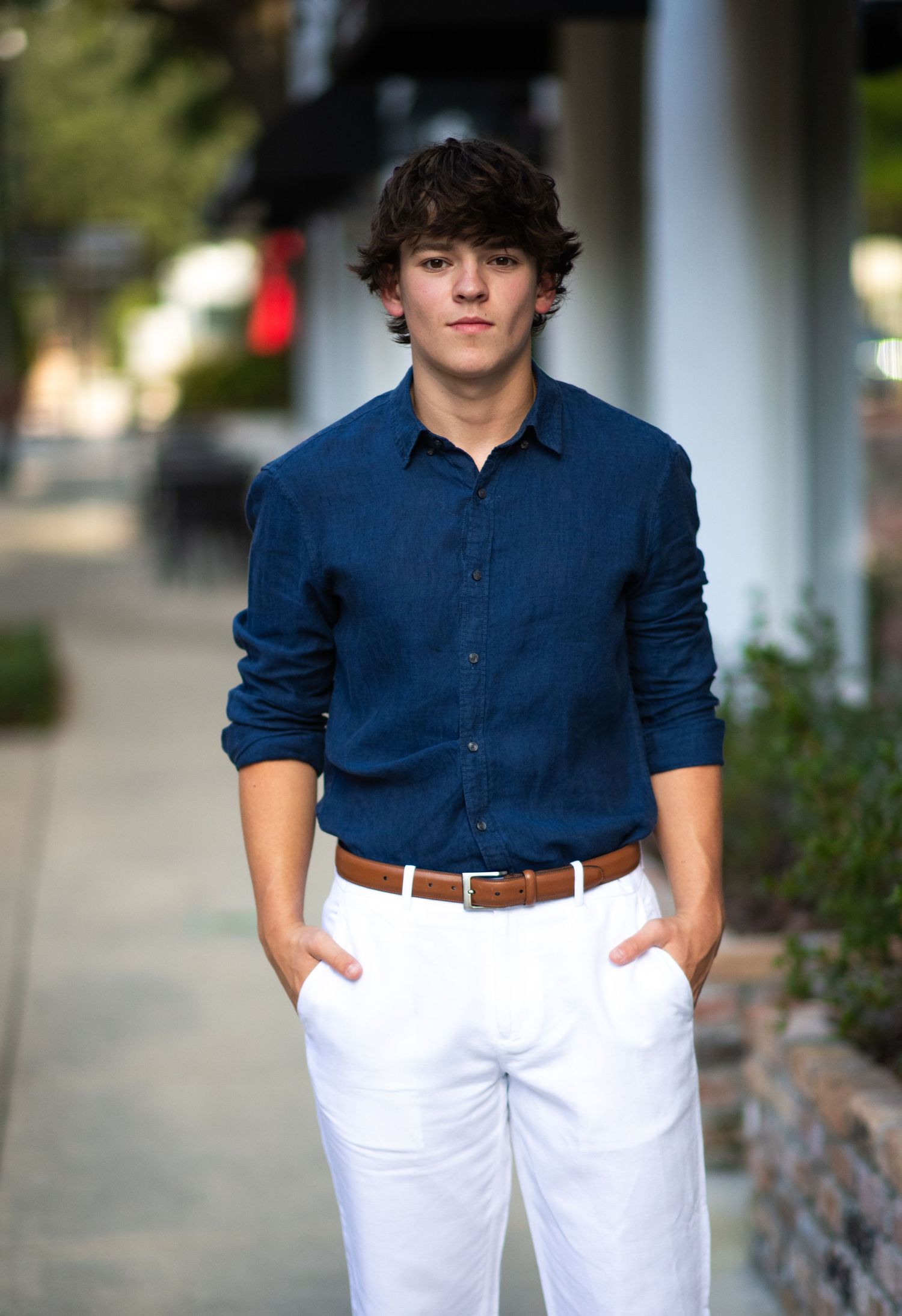 Teen boy in navy shirt and white pants posing on a sidewalk for senior portraits.