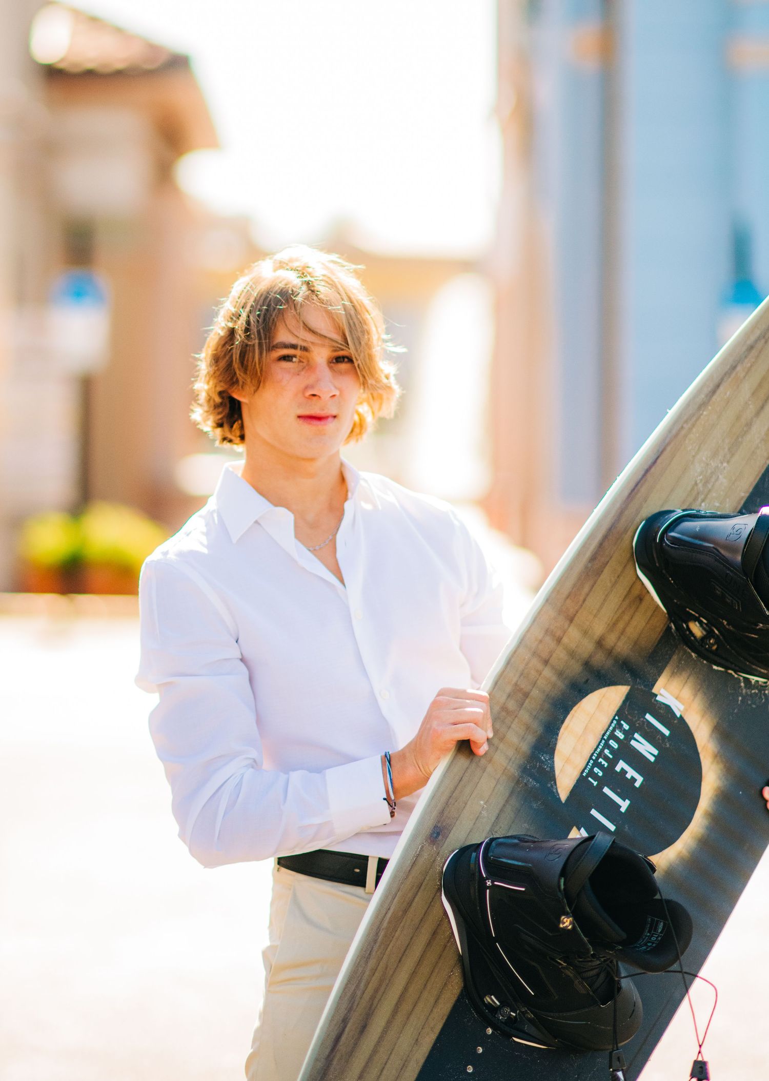Young man in white shirt holding a wakeboard outdoors in a sunny urban setting.