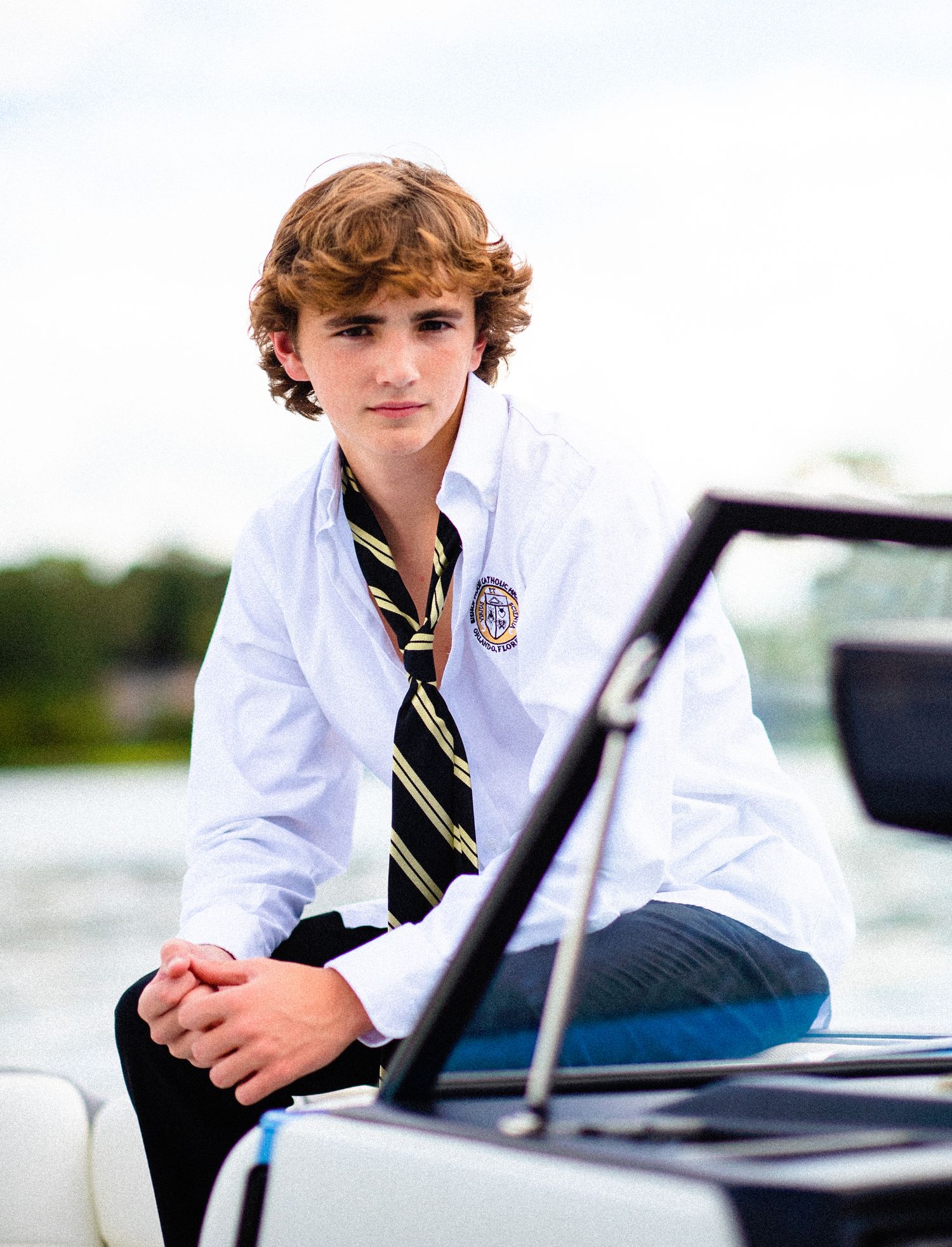 Teen boy in school uniform with striped tie sits on a boat, posing for a portrait on the water.