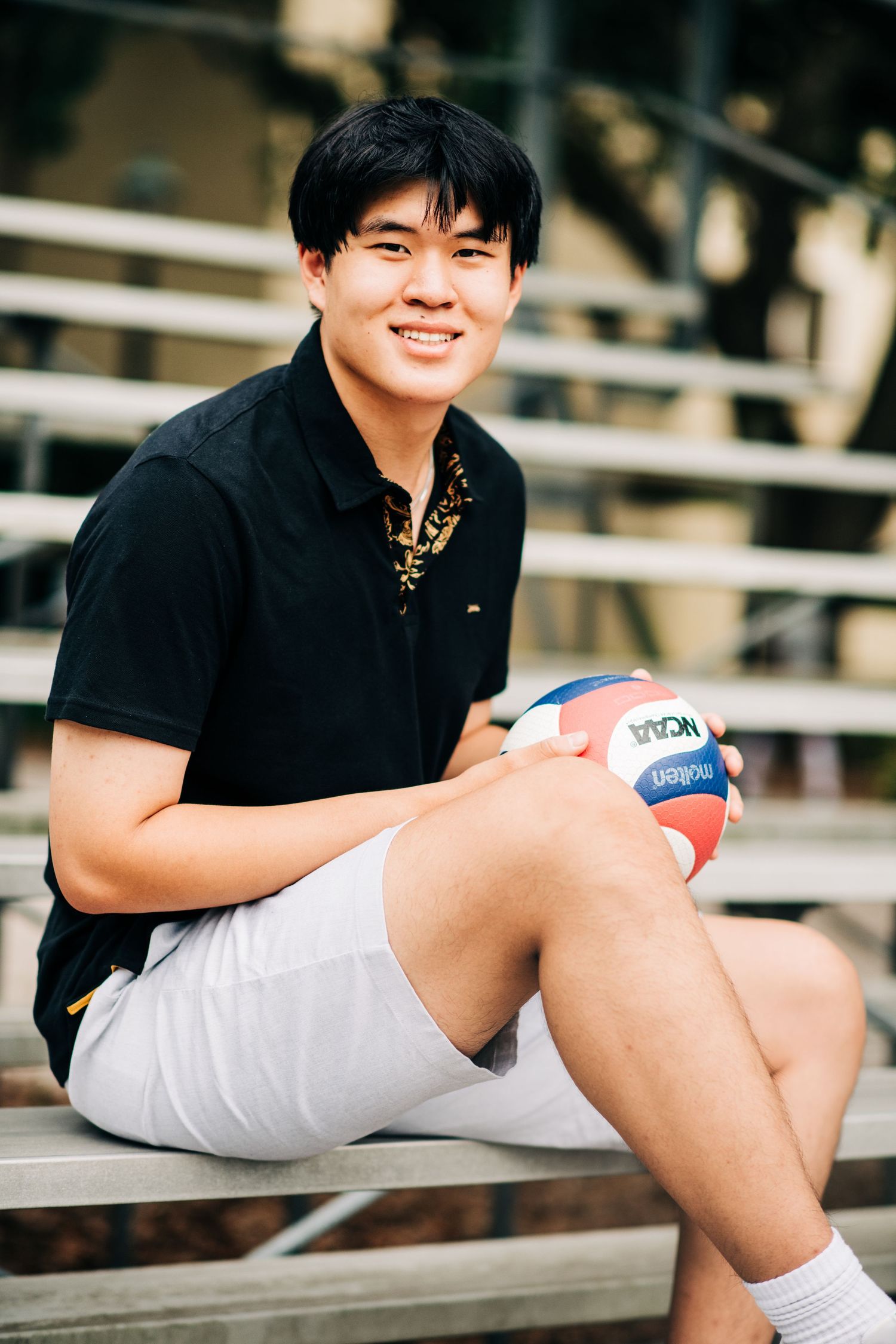 Smiling young man in black polo shirt holding NCAA volleyball while sitting on bleachers outdoors.