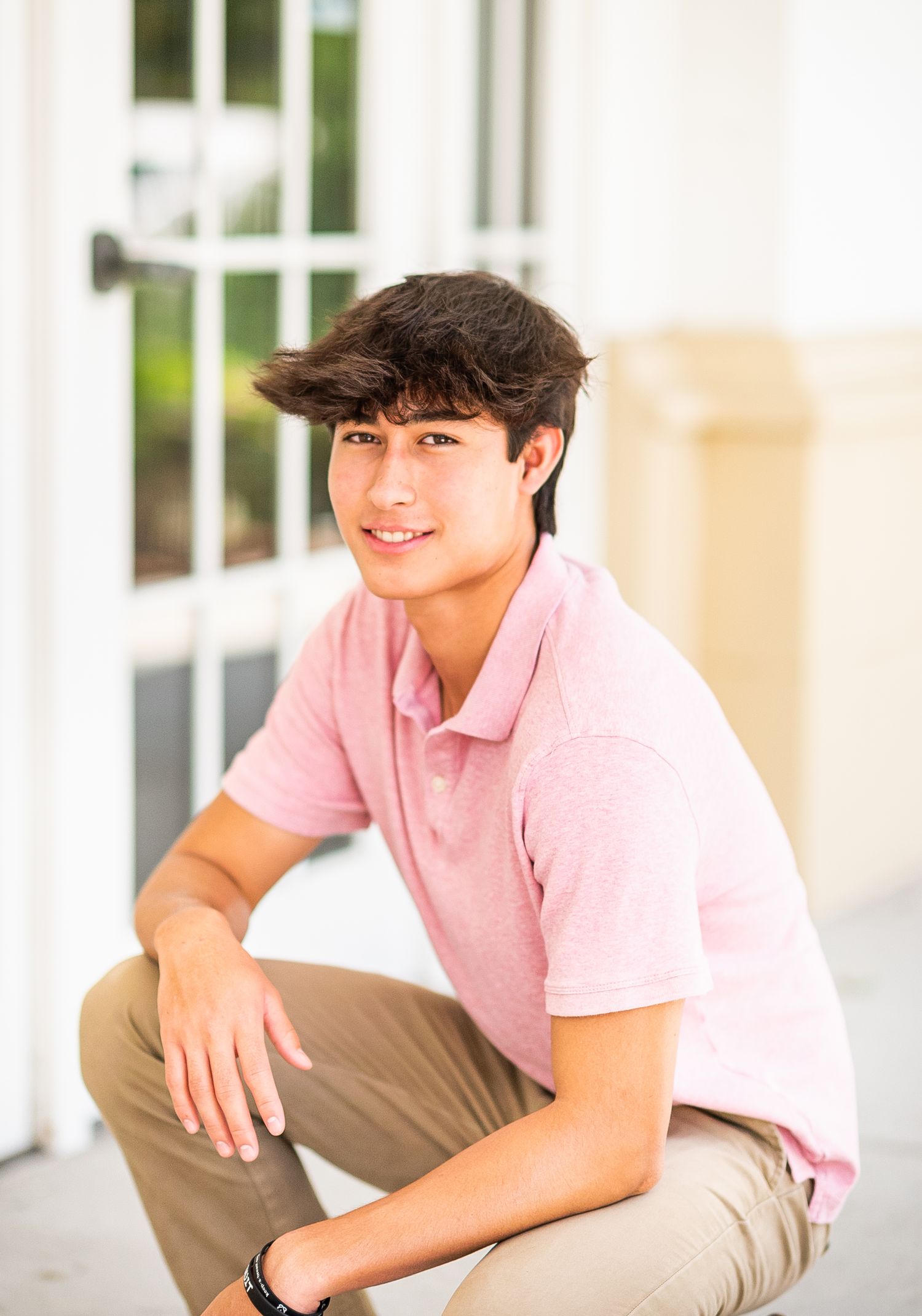 Young teen boy in pink polo shirt and khaki pants crouching outdoors near white French doors, smiling.