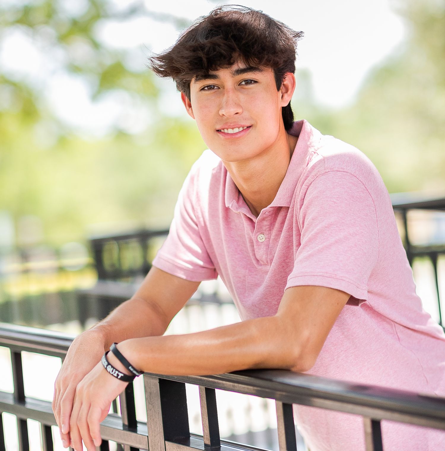 Smiling teenage boy in pink polo shirt leaning on metal railing outdoors in a senior portrait photo.