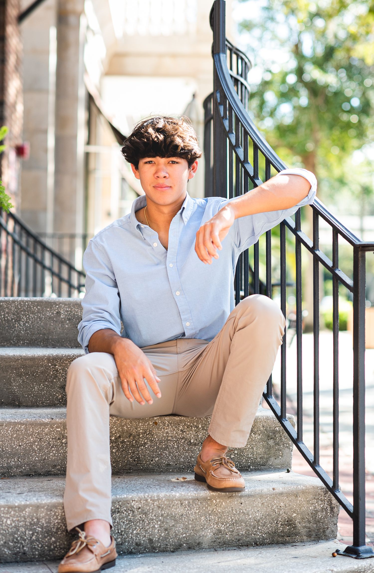 Young man in light blue shirt and khaki pants sitting on outdoor steps leaning on black iron railing.