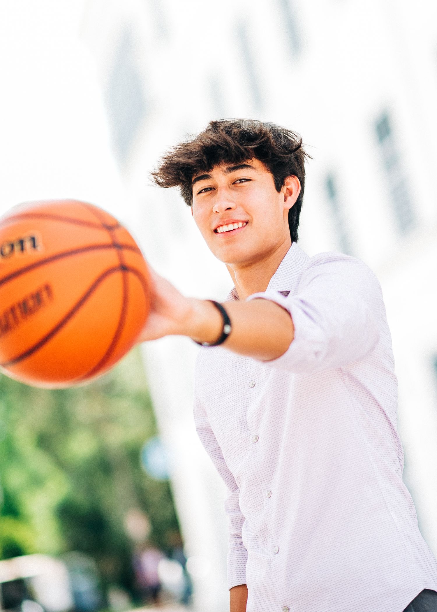 Smiling teenage boy in white shirt holding a basketball outdoors on a sunny day.