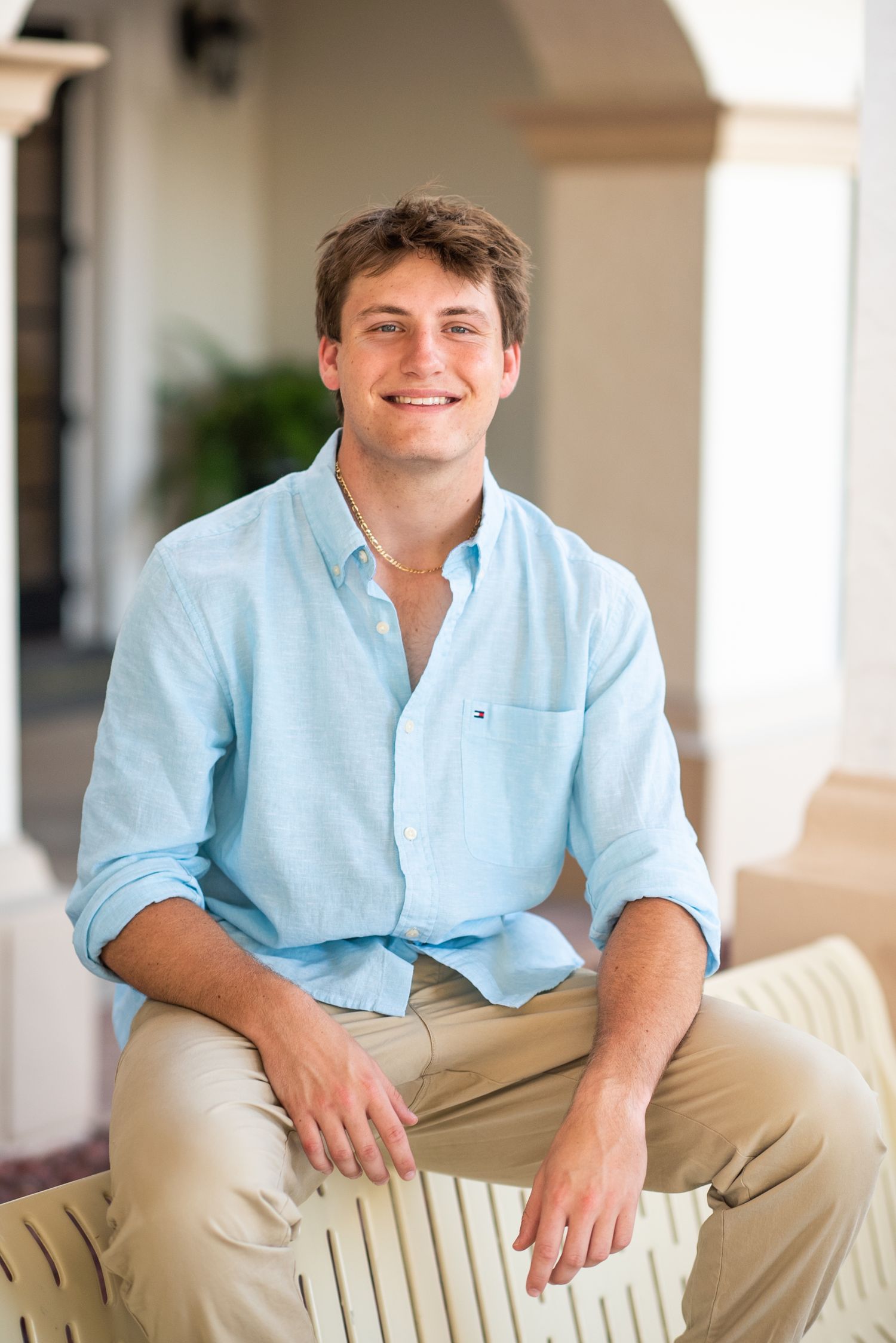 Smiling young man in light blue Tommy Hilfiger shirt sitting on outdoor furniture.