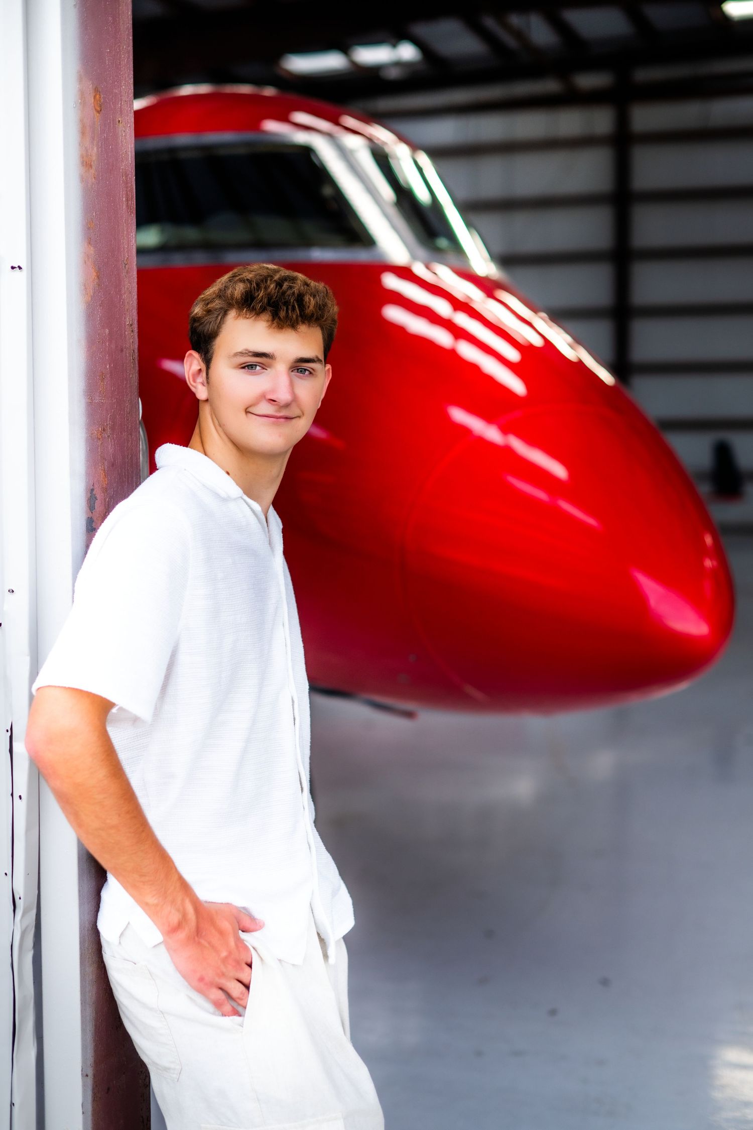 Young man in white outfit leaning against a hangar door with a shiny red aircraft in the background.