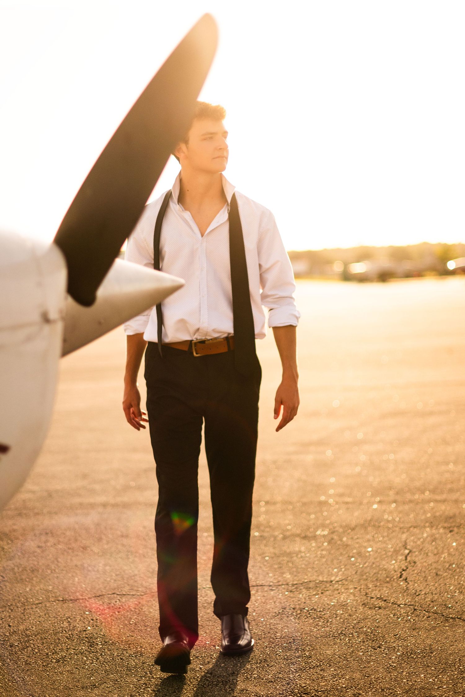 Young man in white shirt and suspenders walks on airport tarmac near propeller plane at golden sunset.