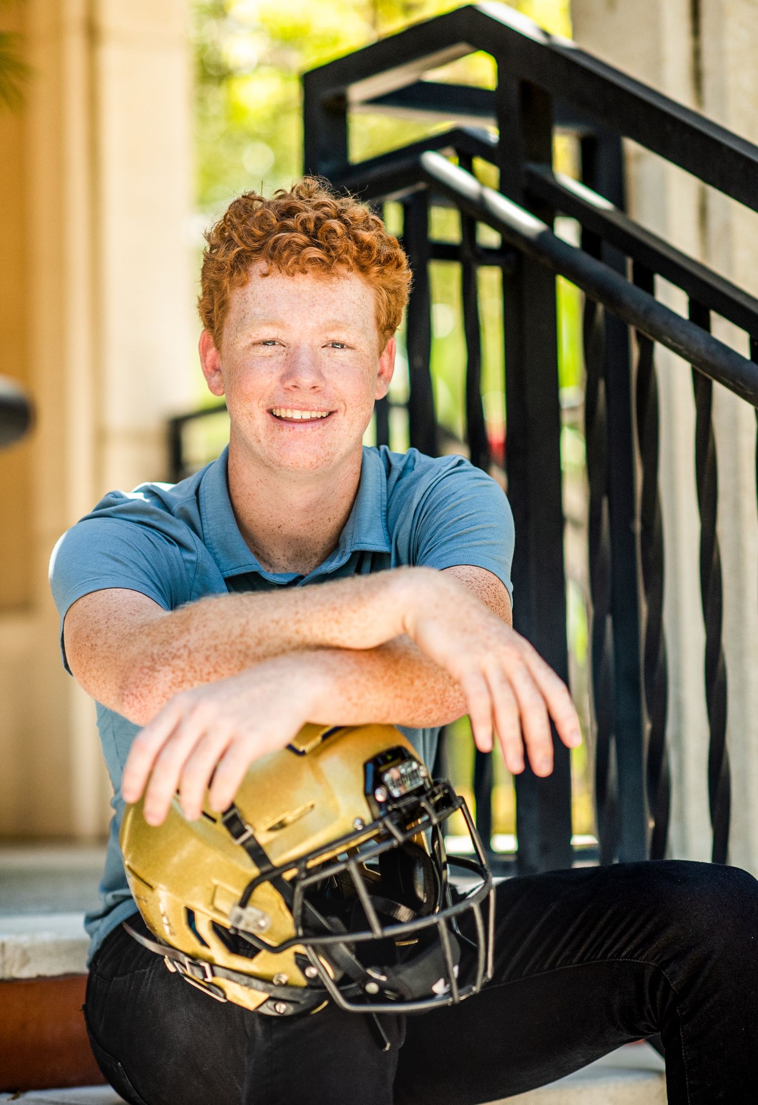 Smiling teenage boy with red hair poses on steps holding a gold football helmet wearing a blue polo shirt.