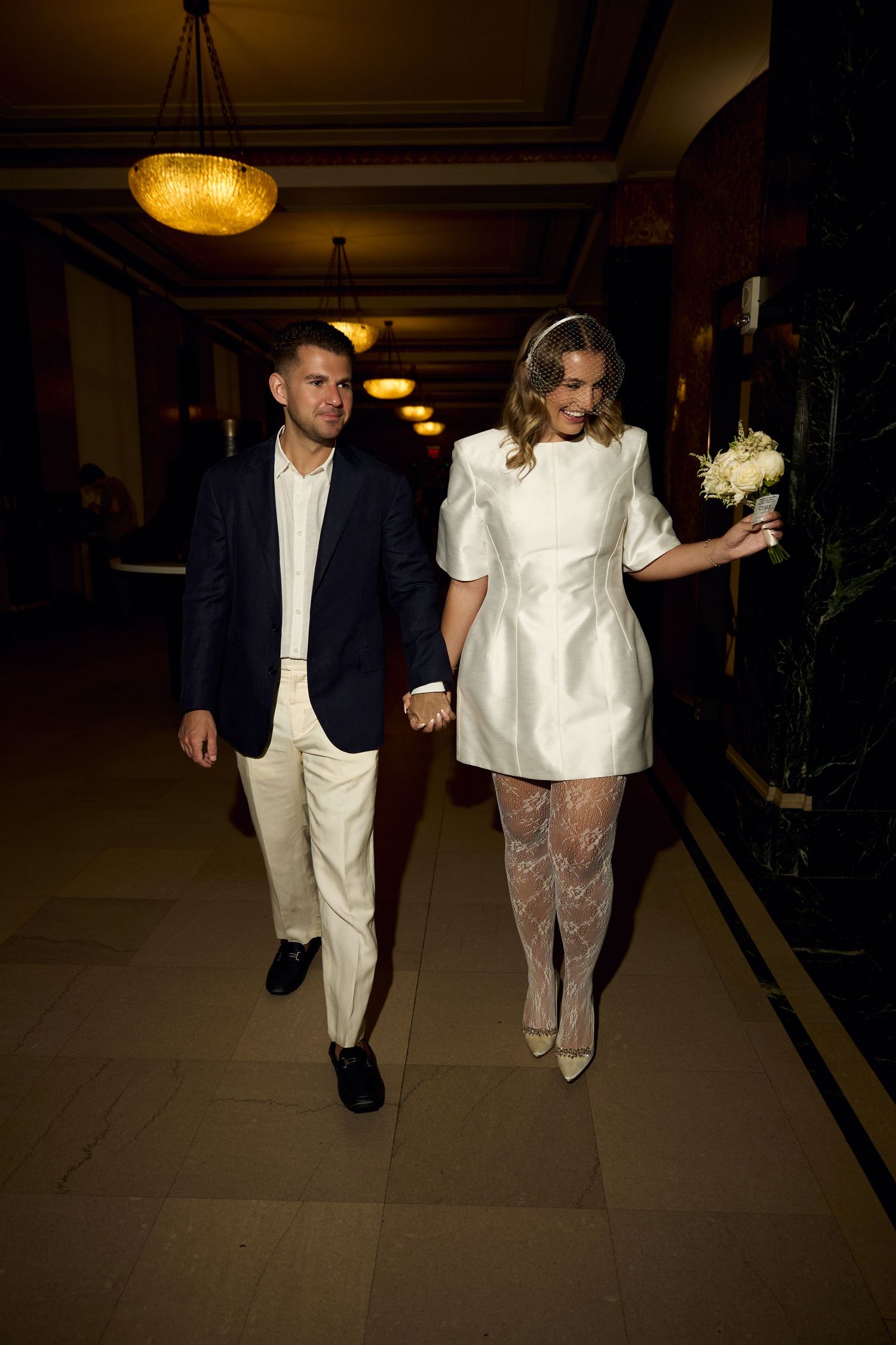 Newlywed couple walking hand in hand, bride in white mini dress with bouquet, groom in navy blazer.
