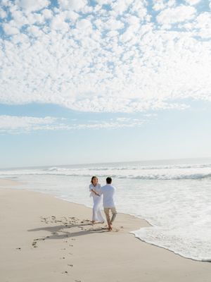 Couple in white dancing on a sandy beach with ocean waves and a beautiful cloudy sky backdrop.