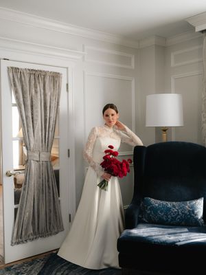 Bride in lace long-sleeve wedding gown holding red bouquet stands by draped window near navy armchair.