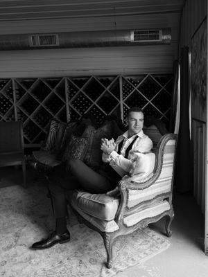 Man in suit sitting relaxed in ornate chair beside wine rack in black and white photo.
