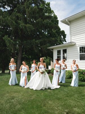 Bride in white ball gown poses with six bridesmaids in light blue strapless gowns on green lawn beside white house.