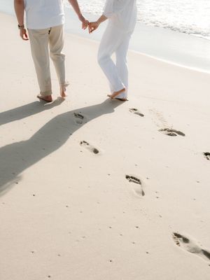 Couple holding hands walking barefoot on white sandy beach, leaving footprints in the sand near ocean waves.