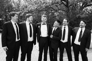 Black and white photo of six men in suits and tuxedos smiling together outdoors at a wedding.