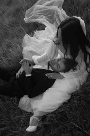 Black and white wedding photo of bride and groom lying in grass field, bride in white gown cradling groom in tuxedo.