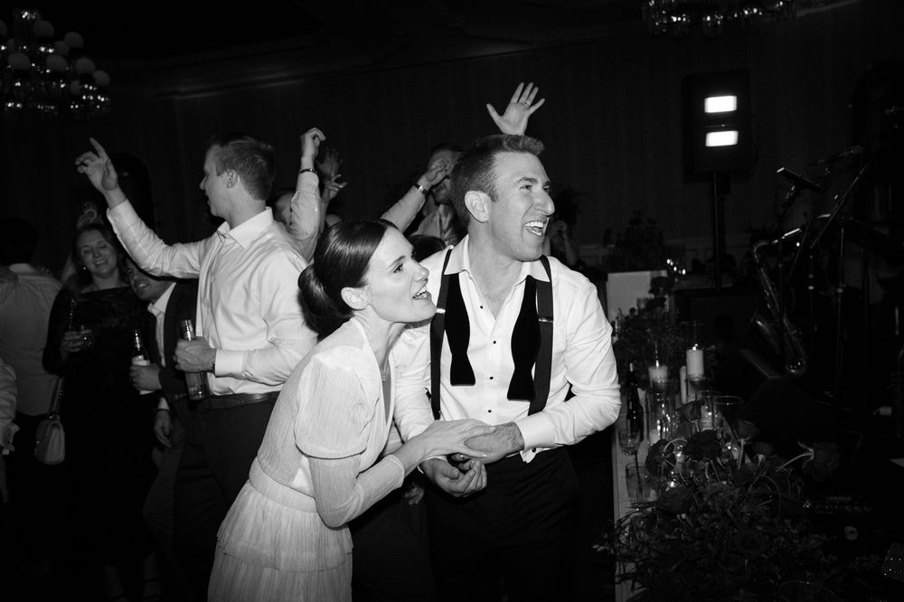 Black and white photo of a couple dancing joyfully at a wedding reception with guests celebrating in the background.