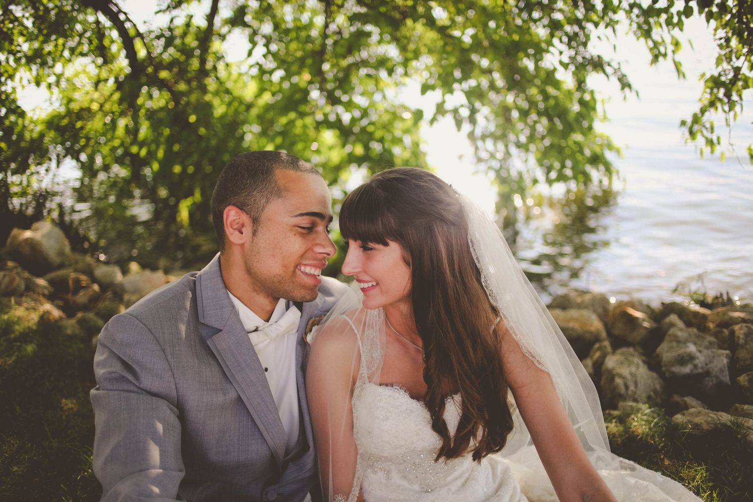 A smiling bride and groom share a romantic moment outdoors near a rocky waterfront surrounded by lush green trees.