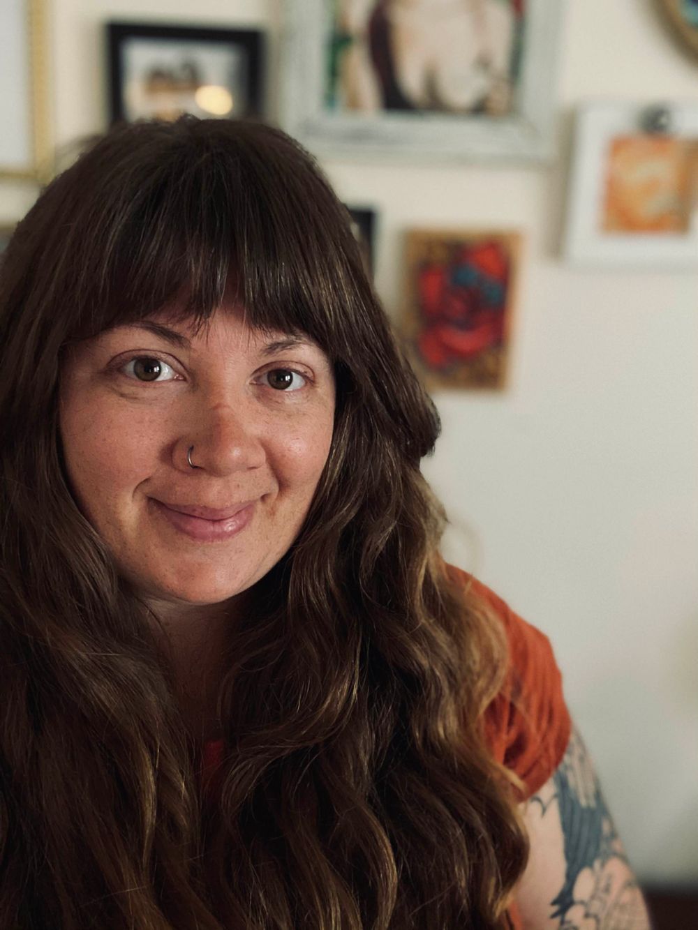 Smiling woman with long wavy brown hair and bangs poses in front of a wall decorated with framed artwork.