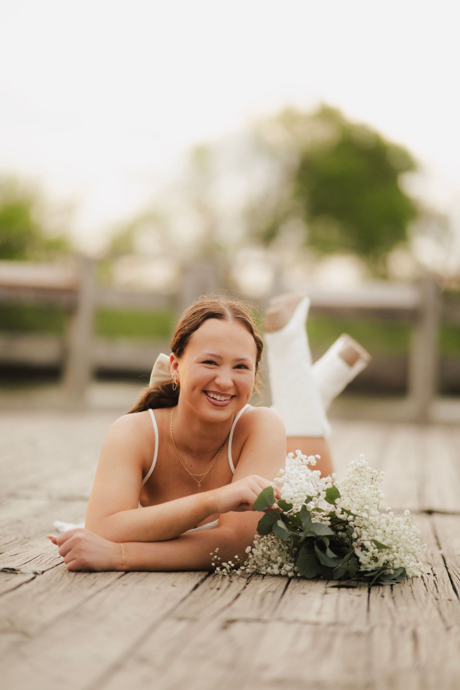 Smiling young woman in white dress lies on a wooden dock holding a bouquet of white wildflowers.