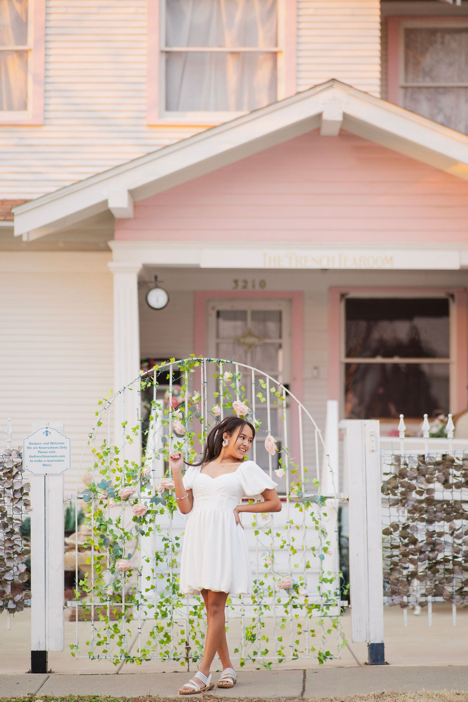 Young woman in white dress stands by a decorative gate in front of a charming pink house at sunset.