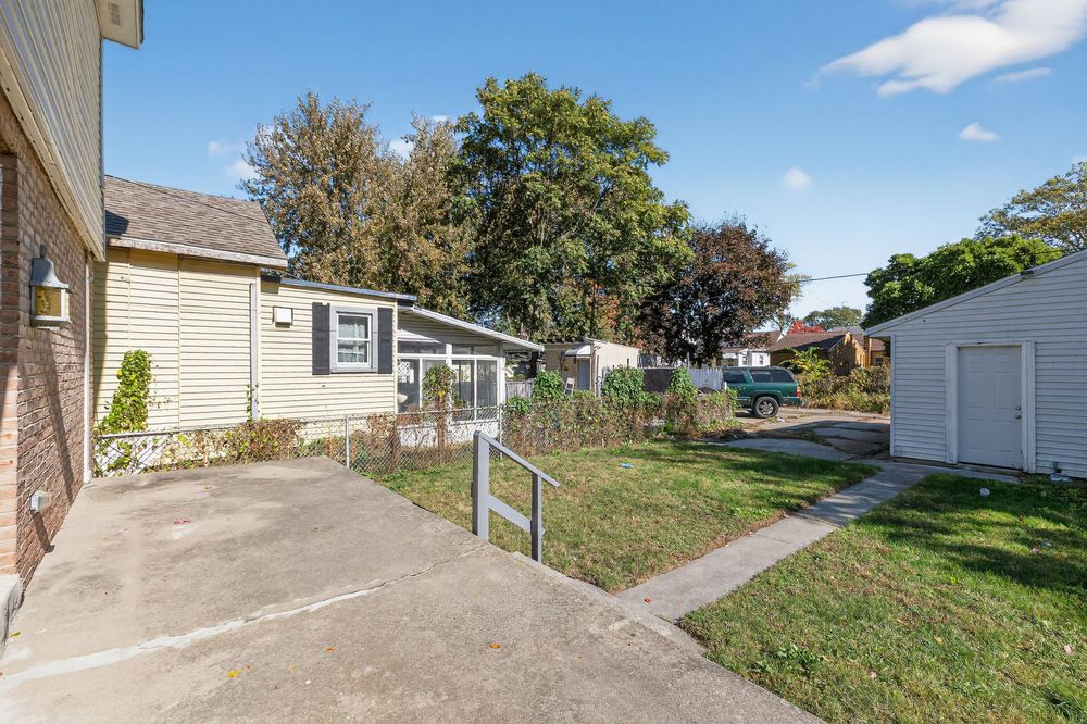 Suburban home exterior with concrete driveway, green lawn, detached garage, and mature trees under a clear blue sky.