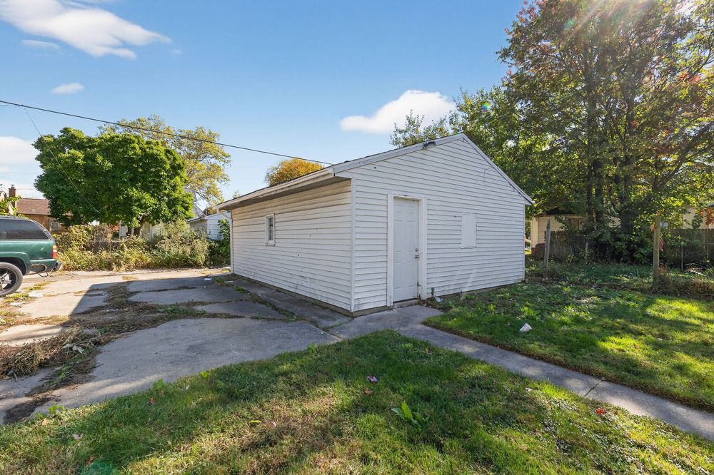 White detached garage with large door sits on a concrete driveway surrounded by green lawn and mature trees on a sunny day.