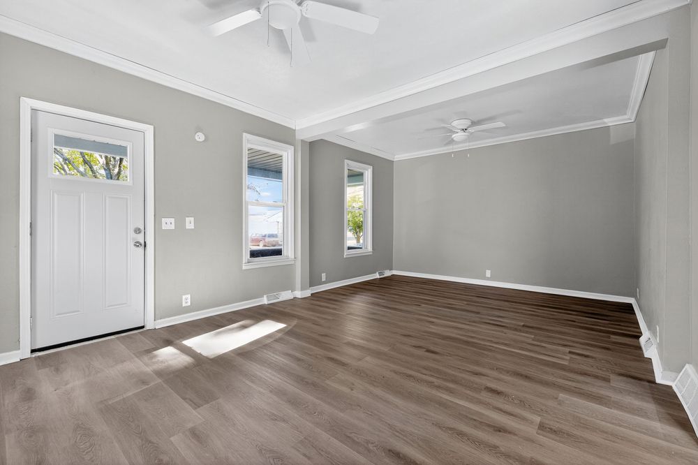 Bright open living room with gray walls, crown molding, ceiling fan, and dark hardwood floors near a white front door.