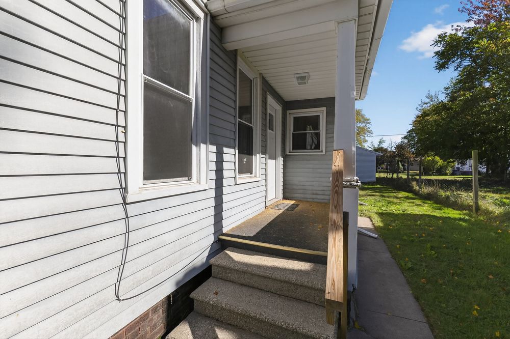 Side entrance of a gray vinyl-sided home featuring wooden steps, a small porch, and a well-maintained green lawn.