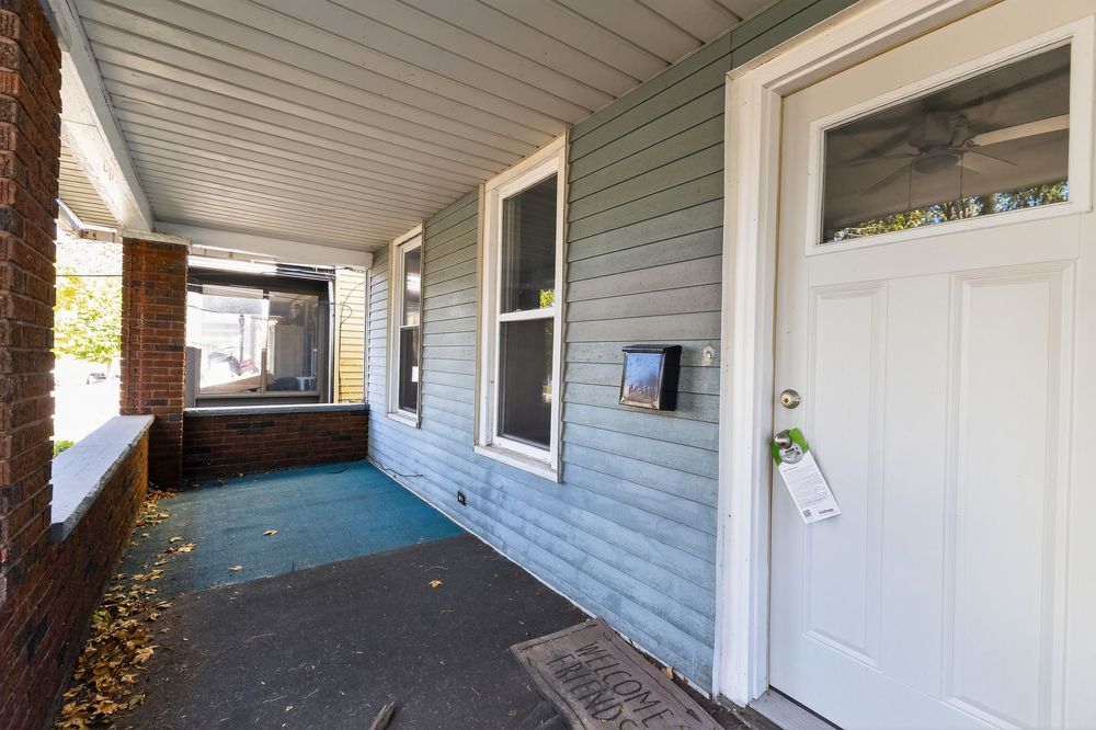 Covered front porch of a house with gray siding, white door, and autumn leaves on the ground.