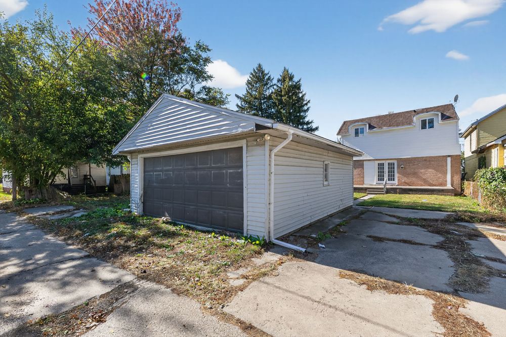 Detached two-car garage with brown doors beside a brick house, surrounded by mature trees and a paved driveway.