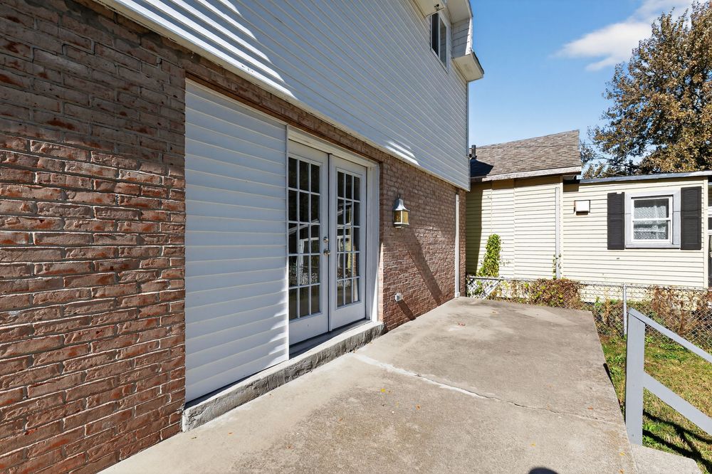 Brick home exterior featuring French doors, concrete patio, and manicured landscaping on a sunny day.