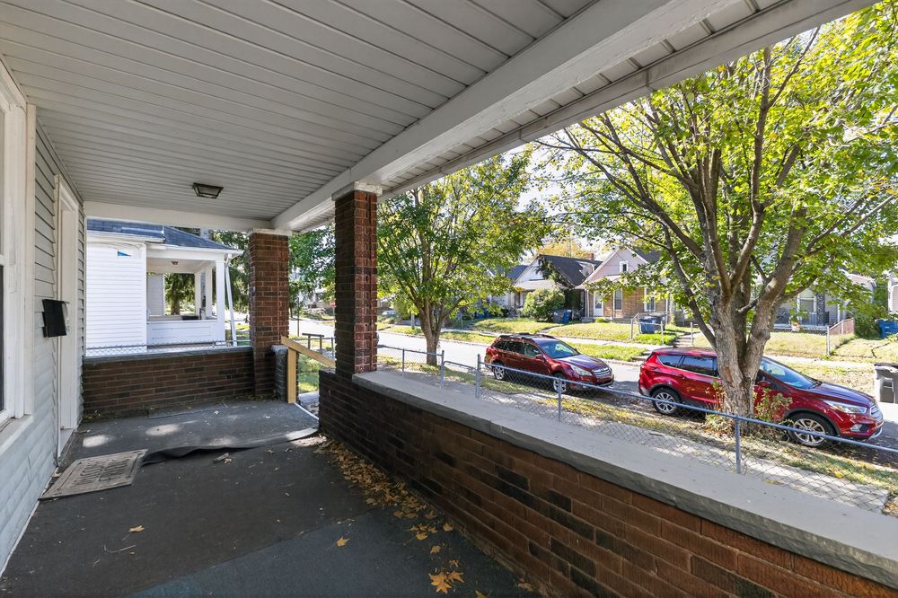 Covered front porch with brick columns overlooking a tree-lined residential street with parked cars on a sunny day.