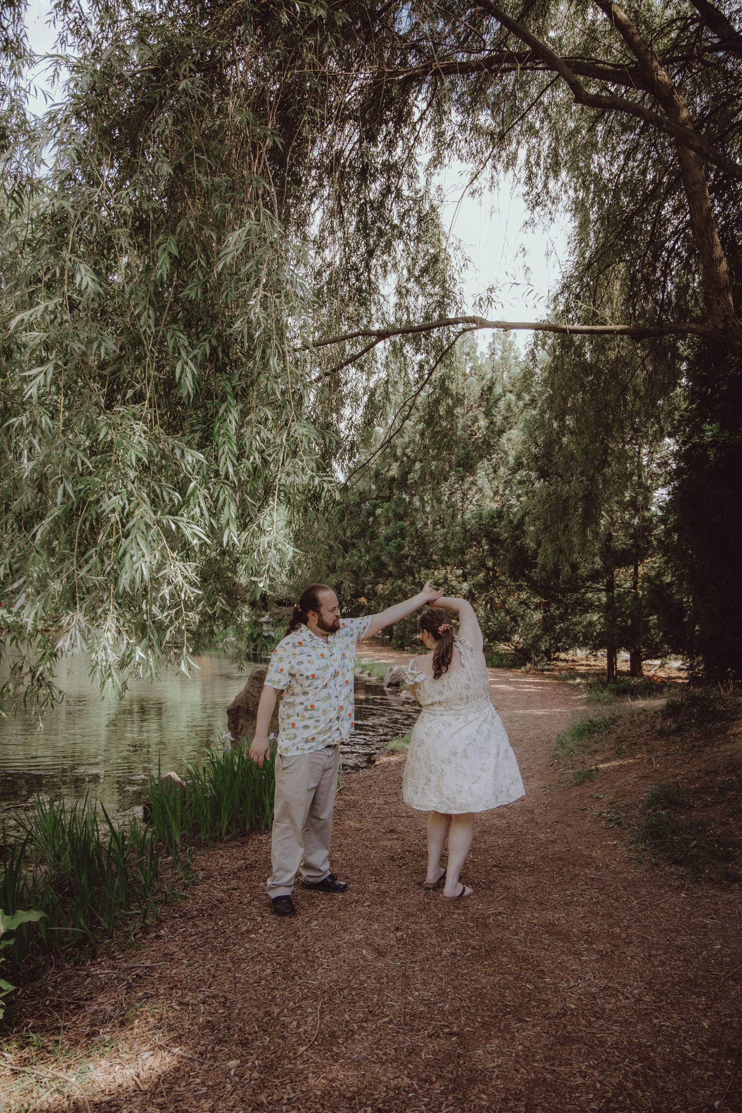 Couple dancing under weeping willow trees by a serene pond, woman in white dress being twirled on a woodland path.