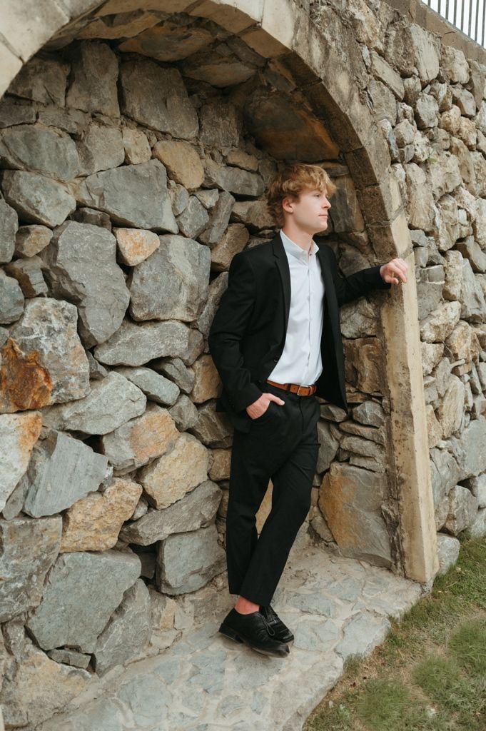 Young man in black suit leaning against stone arch doorway outdoors.