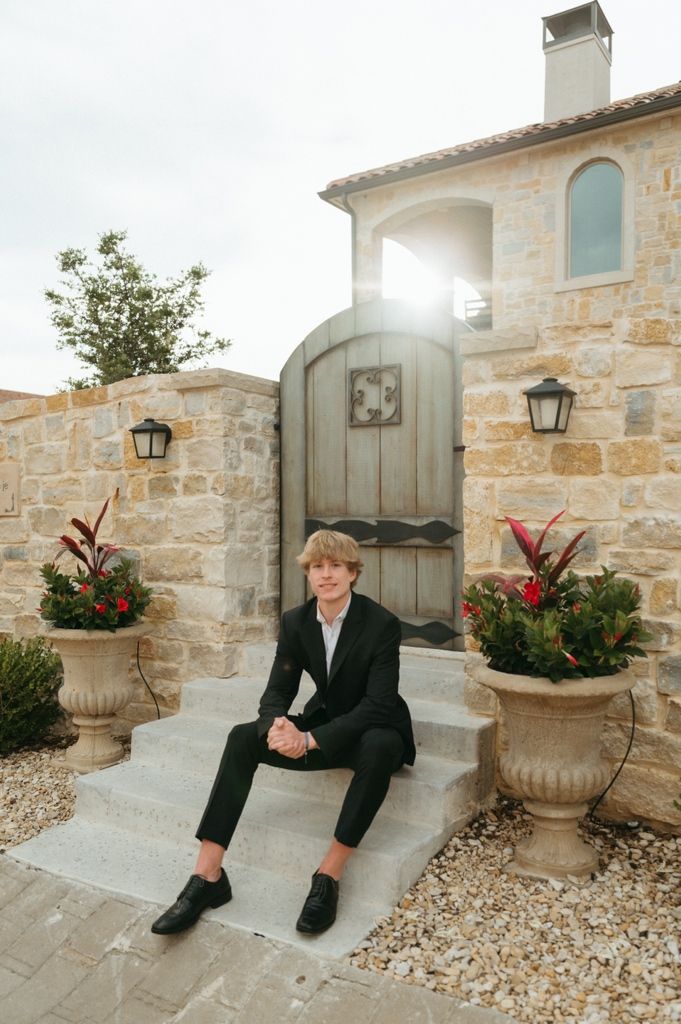 Teen boy in black suit sitting on steps outside stone building with wooden gate.