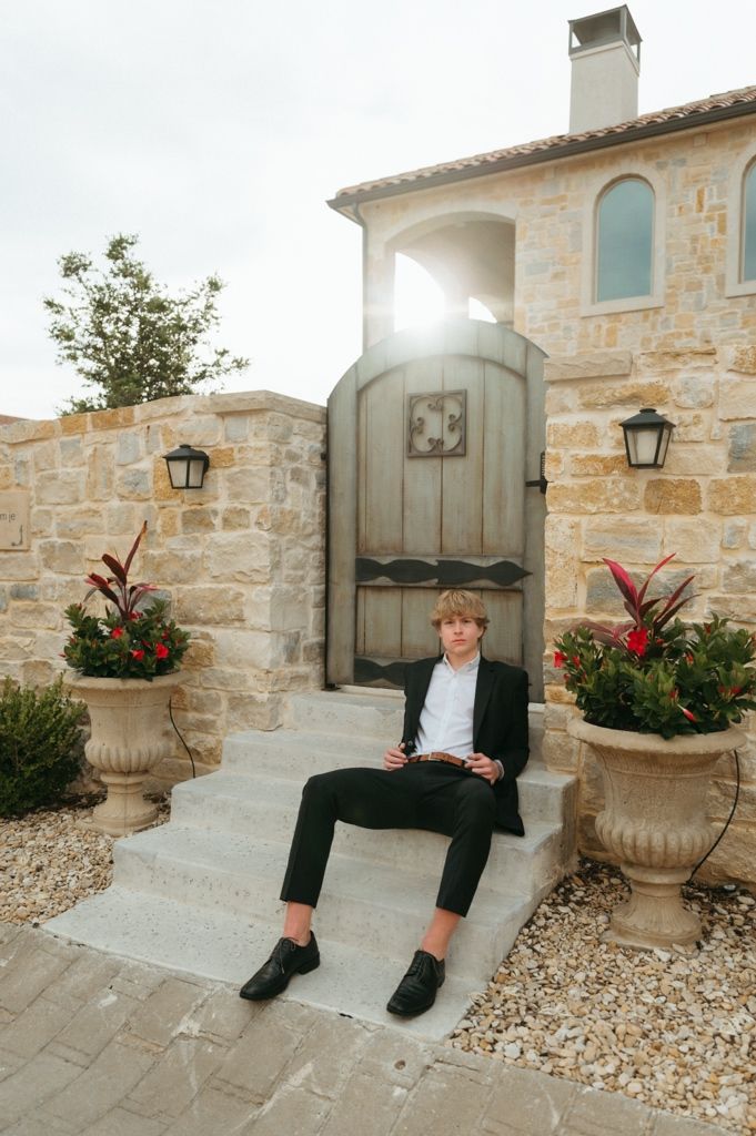 Young man in black suit sitting on stone steps by ornate wooden gate of luxury villa with flower urns.
