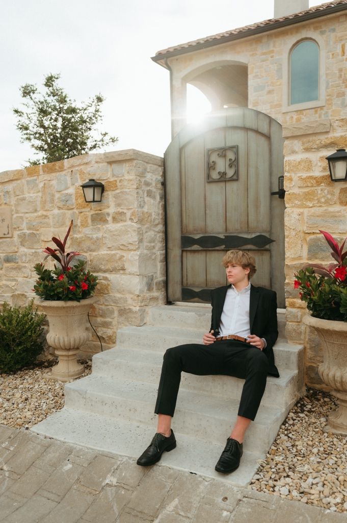 Teenage boy in black suit sits on stone steps by ornate wooden door with sunlight behind.