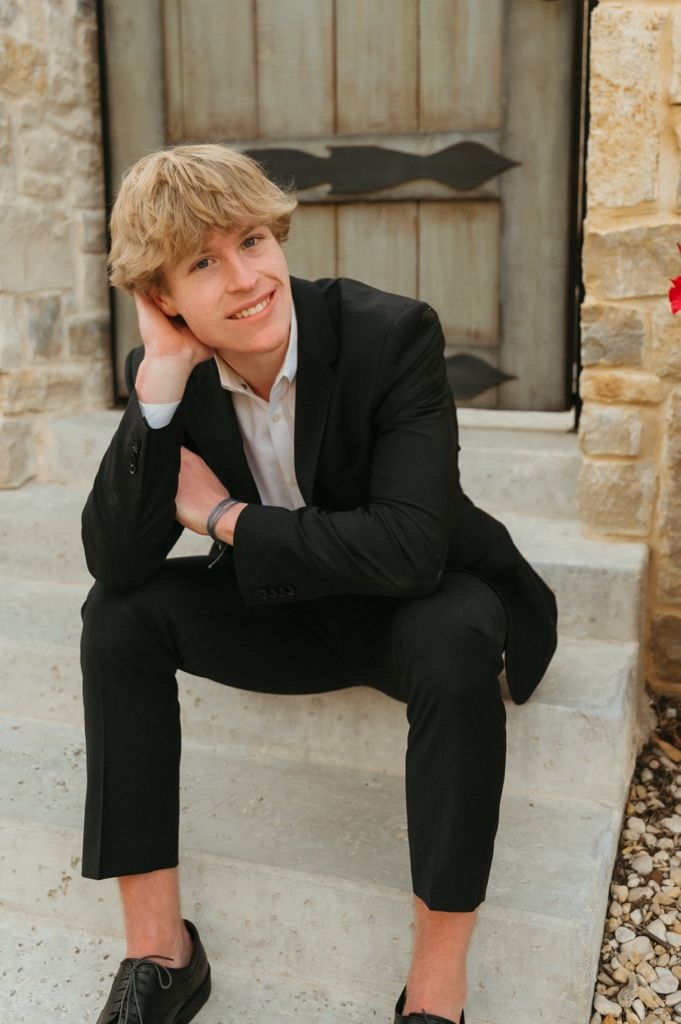 Smiling blonde teen boy in black suit sitting on stone steps outdoors.