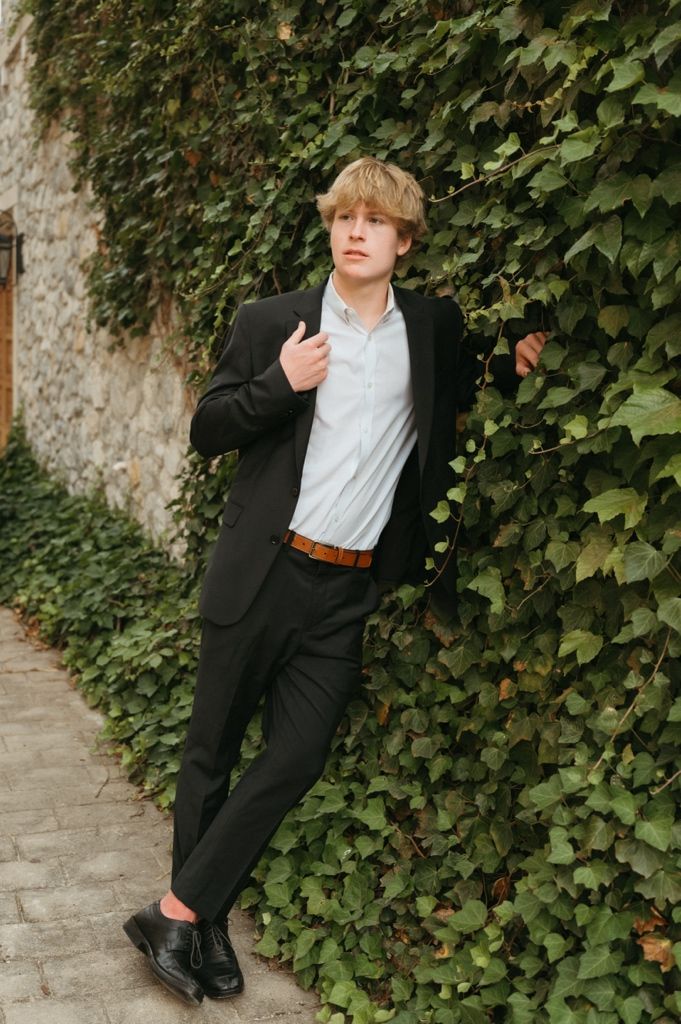 Young man in black suit and white shirt leans against ivy-covered stone wall outdoors.