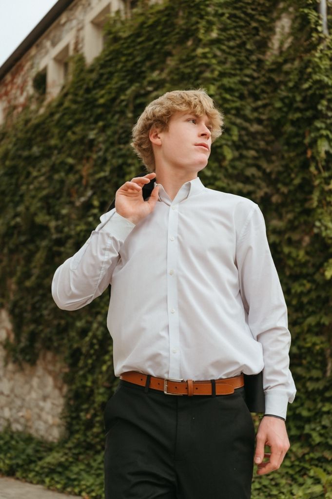 Blond curly-haired young man in white dress shirt walking near ivy-covered stone wall outdoors.