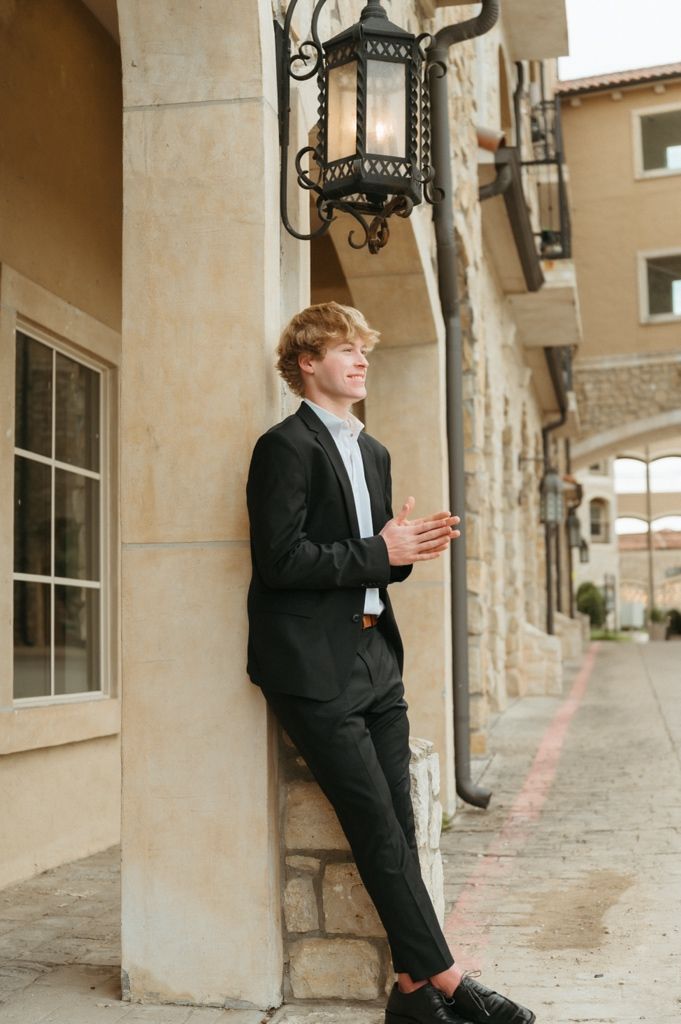 Young man with red hair in black suit leaning against stone pillar under ornate lantern in European-style courtyard.