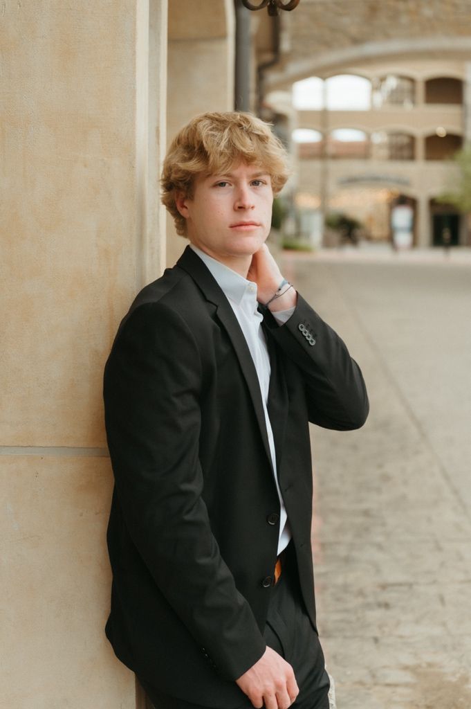 Young man in black suit leaning against stone column, hand on chin, thoughtful pose outdoors.