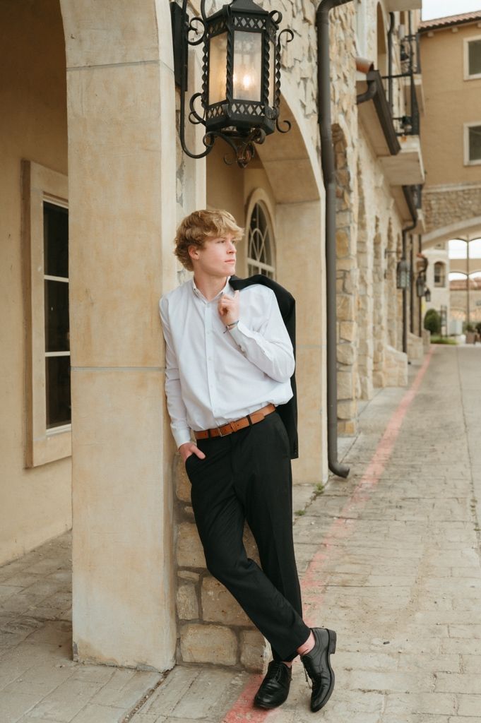 Young blond man in white shirt and black trousers leaning against stone arch column outdoors.