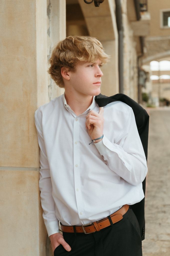 Stylish young man with curly blond hair holding black blazer over shoulder in white dress shirt.