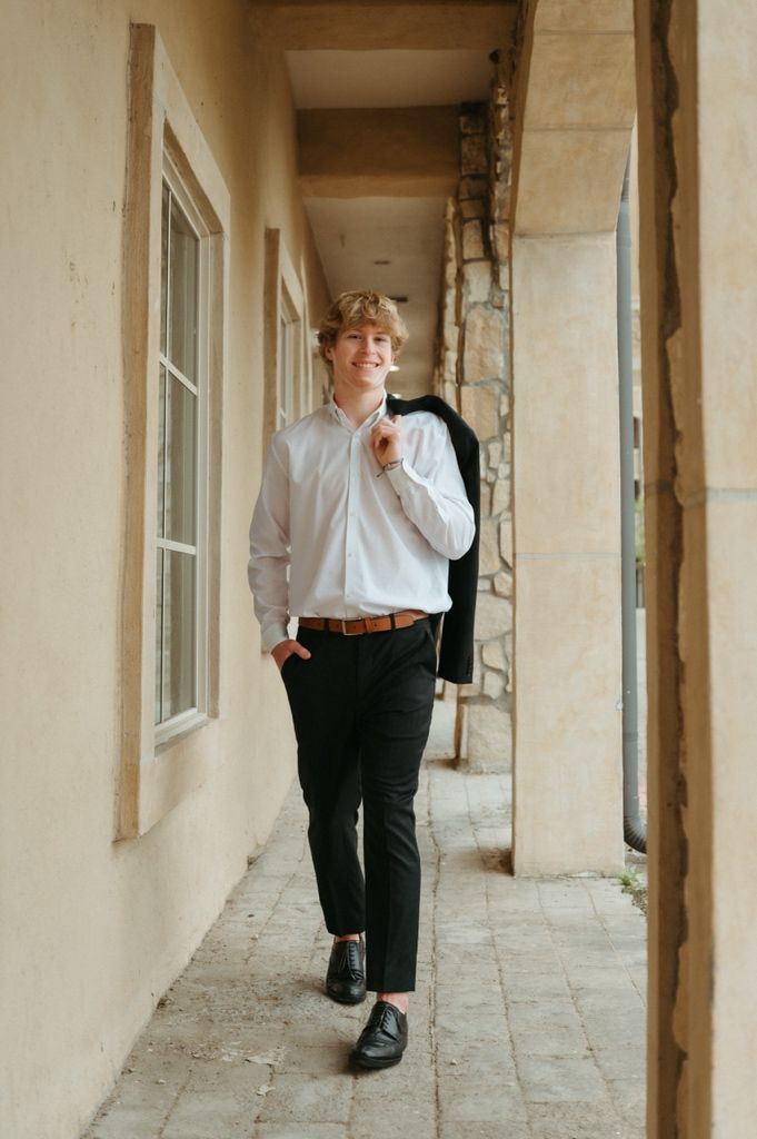 Young man in white shirt and black trousers carrying jacket over shoulder walks along outdoor corridor.