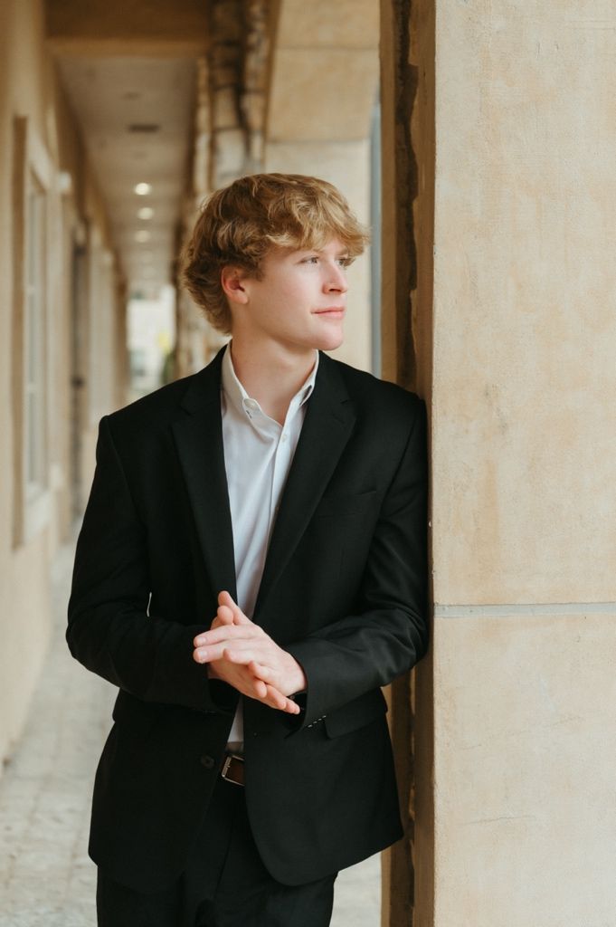 Blonde teen in black blazer leaning on column with clasped hands, gazing away in corridor.